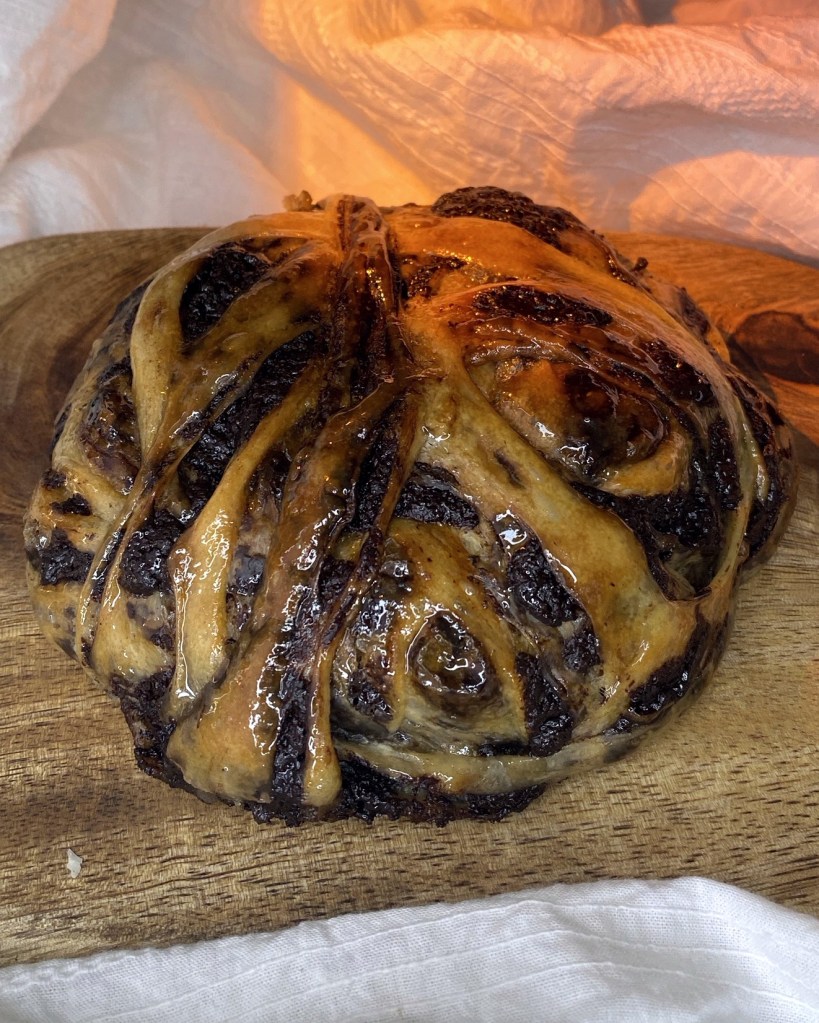 A baked, glossy chocolate bun on a wooden board with strips of dough and chocolate filling twisted around each other.